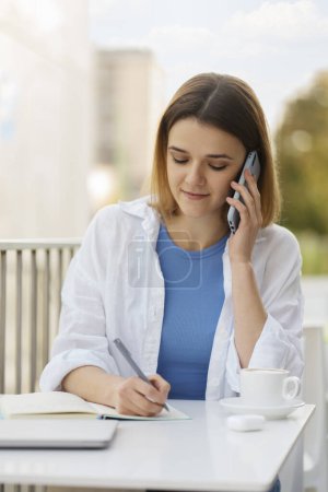 Photo for Portrait of smiling woman talking on mobile phone, taking notes, working project sitting at workplace. Technology concept - Royalty Free Image