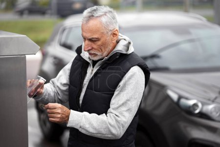 Photo for Elderly gray-haired confident active man, experienced driver inserts coins into a coin acceptor, paying for contactless car wash in self-service machine. Car maintenance Auto service Economy Business - Royalty Free Image