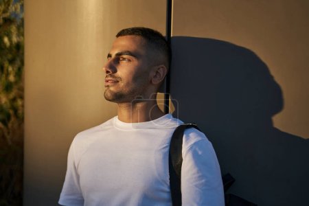 Téléchargez les photos : Jeune bel homme souriant du Moyen-Orient portant un t-shirt blanc regardant loin profitant du coucher de soleil dans la rue - en image libre de droit