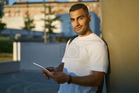 Foto de Young attractive smiling arab man holding digital tablet looking at camera on the street. Portrait of middle eastern student standing in university campus - Imagen libre de derechos