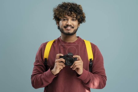 Photo for Smiling Indian photographer holding photo camera taking pictures isolated on blue background. Portrait of happy attractive asian tourist with backpack looking at camera. Travel concept - Royalty Free Image