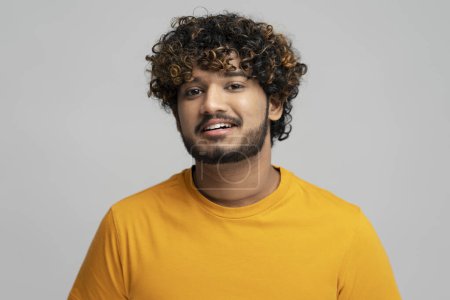 Téléchargez les photos : Portrait d'un homme indien attrayant et souriant aux cheveux bouclés élégants portant un t-shirt jaune isolé sur fond gris. Confiant positif asiatique étudiant regardant caméra, studio shot - en image libre de droit