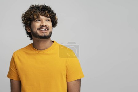 Foto de Retrato de atractivo hombre indio sonriente con el pelo rizado con estilo usando camiseta amarilla, mirando hacia otro lado, espacio de copia. Feliz modelo asiático posando para fotos aisladas sobre fondo gris en estudio - Imagen libre de derechos