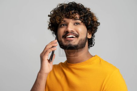 Foto de Retrato de primer plano de hombre indio sonriente confiado hablando en el teléfono móvil mirando hacia otro lado aislado sobre fondo gris. Feliz hipster asiático moderno respondiendo a la llamada. Concepto tecnológico - Imagen libre de derechos