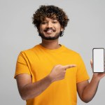 Attractive smiling Indian man holding mobile phone, shopping online pointing finger at empty display isolated on gray background. Modern happy asian hipster using mobile app, ordering food. Mockup