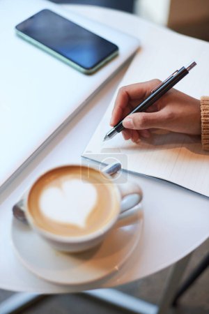 Photo for Selective focus of the hand of a woman holding an ink pen and writing on a white paper sheet, sitting at a table while a coffee break. Mobile phone with empty screen with copy space on office table - Royalty Free Image