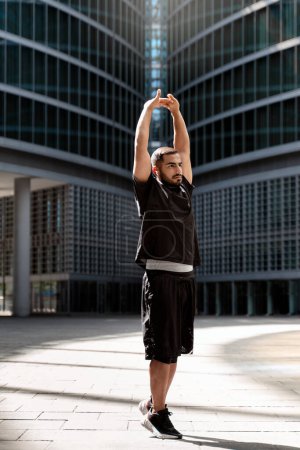 Téléchargez les photos : Jeune homme iranien portant des vêtements de sport échauffant en plein air. Homme sportif étirant le bras avant de courir sous le bâtiment. Entraînement athlète travailler concept - en image libre de droit