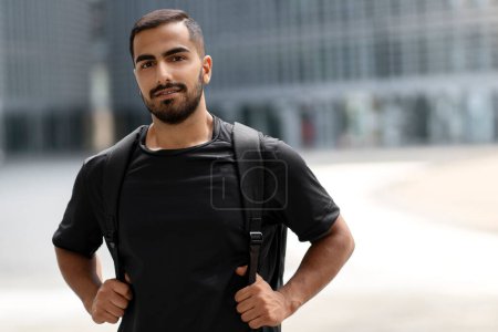 Téléchargez les photos : Happy handsome bearded student looking at camera and smiling while posing with backpack outdoors alone. Positive lifestyle concept - en image libre de droit