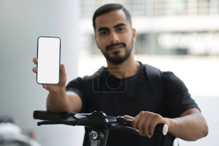 Téléchargez les photos : Heureux homme d'affaires réussi regardant la caméra et montrant smartphone avec écran vide tout en se tenant debout avec scooter électrique - en image libre de droit