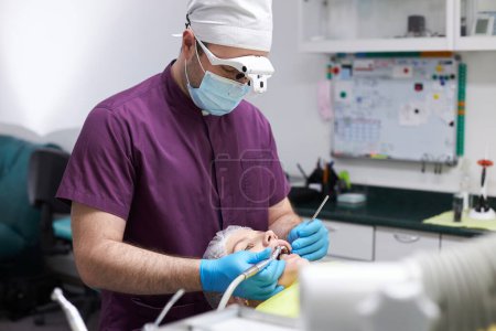 Photo for Male dentist orthodontist hygienist, in medical protective face mask and uniform, using dental equipment and mirror, examining oral cavity and curing caries of a young female patient at dental clinic - Royalty Free Image