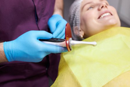 Photo for Selective focus on a modern dental equipment- an ultraviolet lamp for tooth filling in dentist's hand over a blurred smiling female patient, lying on the dentist chair during a dental teeth treatment - Royalty Free Image