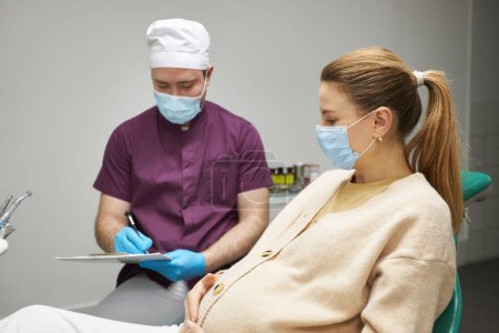 Photo for Caucasian young pregnant woman, wearing medical mask, at dentist appointment in dentistry clinic. The concept of a preventive examination by a dentist during pregnancy - Royalty Free Image