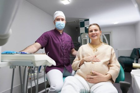Photo for Male dentist doctor and beautiful pregnant woman smiling a cheerful toothy smile, looking at camera after regular dental check-up or teeth treatment in the dental office. People. Service. Healthcare - Royalty Free Image