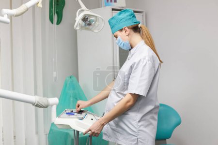 Téléchargez les photos : Side portrait of a young Caucasian pregnant woman, wearing white medical gown and protective mask, a dentist's assistant preparing the workplace in dental office, placing a tray with sterile tools - en image libre de droit