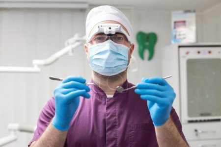 Photo for Confident dental practitioner, dentist, stomatologist orthodontist, wearing medical mask and uniform, holding stainless steel sterile dental probe and mirror for check-up in hands, looking at camera - Royalty Free Image