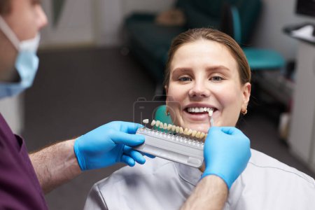 Téléchargez les photos : Gros plan jolie patiente souriante dans le siège du dentiste, regardant la caméra pendant que le médecin orthodontiste, choisissant la nuance de placage dentaire selon le nuancier. La dentisterie esthétique. Pratique dentaire - en image libre de droit