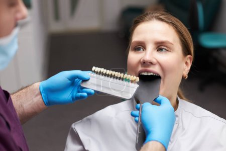 Téléchargez les photos : Close-up woman's face, smiling a toothy smile while a doctor dentist orthodontist, using a tooth color chart, choosing the shade of teeth according to Vita scale. Selection of the color of veneers - en image libre de droit