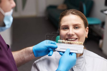 Téléchargez les photos : Médecin dentiste plaçant un nuancier de dents, sur les dents d'une belle femme souriante, en choisissant la nuance de placages, selon l'échelle de Vita. La méthode visuelle de perception subjective de la couleur des dents - en image libre de droit