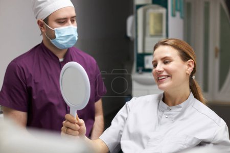Photo for Close-up smiling woman patient in dentist's seat, looking at small mirror while a dentist appointemnt in modern dental clinic. Aesthetic dentistry. Dental practice - Royalty Free Image