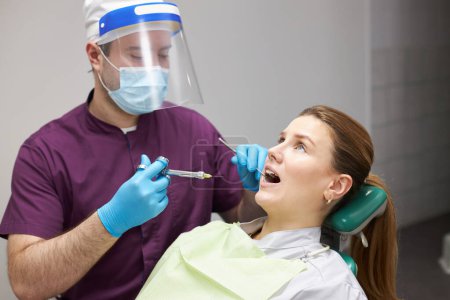 Foto de Caucasian young pregnant woman with open mouth, receiving an injection of dental anesthesia, done by a dentist doctor during teeth treatment in a dentistry clinic. Healthcare and medicine concept. - Imagen libre de derechos