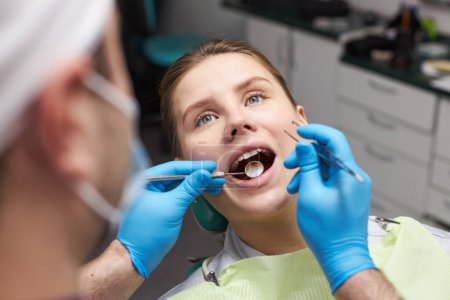 Photo for Close-up of young woman with open mouth at dental check-up. Doctor dentist using dental mirror and sterile stainless steel probe, examining teeth and gum of female patient. Dental practice. Dentistry - Royalty Free Image
