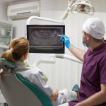 Rear view of an orthodontist dentist pointing at a digital x-ray orthopantomogram, explaining to a female patient the treatment that needs to be done in a dental clinic. Dentistry. Dental practice