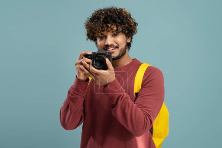 Foto de Smiling curly haired Asian guy photographer, journalist or traveler with yellow backpack, holding digital camera and aiming looking at camera, before taking photos, over isolated blue color background - Imagen libre de derechos