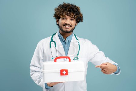 Photo for Handsome confident young Asian male doctor, physician, in white medical coat, pointing with index finger at first aid kit, smiling a toothy smile, looking at camera isolated on blue color background - Royalty Free Image