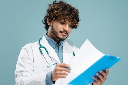 Photo for Young Indian male doctor in white medical coat, with stethoscope, holding clipboard, examining a patient's medical history, isolated on blue background. Medical insurance, healthcare, medicine concept - Royalty Free Image