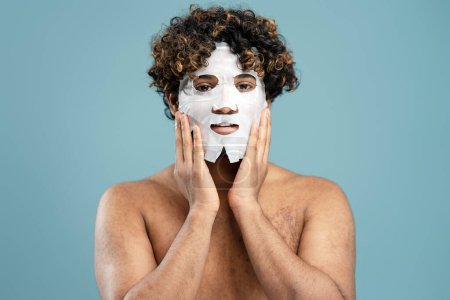 Photo for Shirtless curly-haired young Hindu man applying on his face a smoothing moisturizing textile tissue mask, looking at camera isolated over a blue background. Male beauty and skin care concept - Royalty Free Image