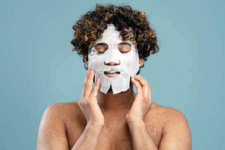 Téléchargez les photos : Male beauty concept. Curly-haired ethnic naked young manapplying tissue fabric textile mask on his face, isolated on blue color background. Skin care, home spa and cosmetic procedures concept. Closeup - en image libre de droit