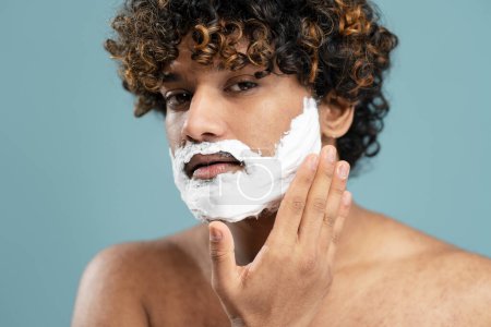 Foto de Retrato de belleza masculino de cerca de un joven indio sin camisa de pelo rizado, tocándose la barba mientras se aplica espuma de afeitar en la cara sobre un fondo azul aislado. Concepto de cuidado de la piel y el cuerpo - Imagen libre de derechos