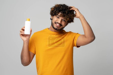 Téléchargez les photos : Homme indien attrayant souriant tenant une bouteille blanche de shampooing hydratant nourrissant anti pellicules, regardant la caméra sur fond gris isolé. Soins capillaires et traitement médical de la perte de cheveux - en image libre de droit