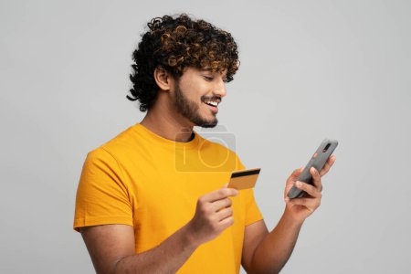 Téléchargez les photos : Handsome smiling Asian ethnic bearded man holding credit card and using mobile phone, inserting data while making online transactions, transferring money via internet banking, isolated gray background - en image libre de droit