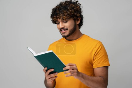 Foto de Confident young Indian man entrepreneur freelancer in yelow t-shirt, holding a notebook, reading his plans or list-to-do, isolated on gray background. Inspired Asian guy reading book - Imagen libre de derechos