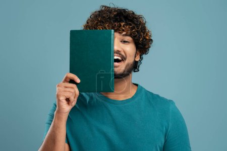 Téléchargez les photos : Happy confident young Indian male student in blue casual t-shirt, smiles looking at camera, covers half of his face with a green hardcover book, isolated on blue background. People Education Business - en image libre de droit