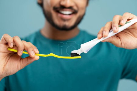 Foto de Close-up of a blurred bearded Indian man in blue t-shirt, smiling a toothy smile while squeezing out toothpaste on a toothbrush, isolated on blue color background. Dental care and oral hygiene concept - Imagen libre de derechos