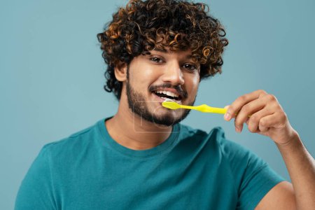 Photo for Smiling happy young Asian man brushing teeth and looking at camera, isolated on blue background. Studio shot of a handsome Indian guy during daily morning hygiene for healthy teeth and beautiful smile - Royalty Free Image