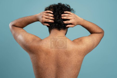 Téléchargez les photos : Young shirtless dark-haired curly man scratching head or massaging head skin standing back to the camera, isolated on blue background. Skin, body and hair care concept. Male beauty portrait - en image libre de droit