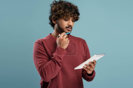 Foto de Confident thoughtful young Hindu man looking at the notepad, holding a pen at his chin while writing the list to do over isolated blue background. Portrait of a businessman, male entrepreneur, student - Imagen libre de derechos