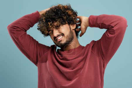 Téléchargez les photos : Young curly-haired bearded Indian man scratching his head on isolated blue background. Pediculosis. Annoying itch. Man with lice and nits. Concept with Healthcare And Medicine. - en image libre de droit