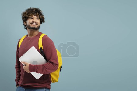 Foto de Young Asian man, smiling student in casual clothes, yellow backpack holding laptop PC, isolated on blue wall background. Concept of education in high school, university or college. Mock up. Copy space - Imagen libre de derechos