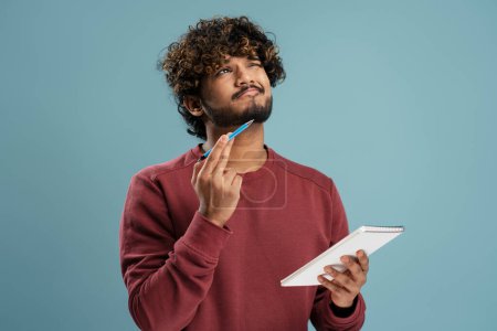 Foto de Pensive young Indian man, businessman, freelancer, entrepreneur holding a notepad and dreamily looking up thinking on new business project, writing list-to-do, isolated on blue color background - Imagen libre de derechos