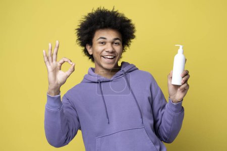 Téléchargez les photos : Jeune homme afro-américain souriant tenant bouteille de shampooing capillaire, montrant ok signe regardant la caméra isolée sur fond jaune, plan studio. Soins capillaires, concept de routine matinale - en image libre de droit