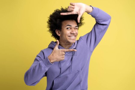 Foto de Joven fotógrafo afroamericano sonriente haciendo marco con las manos mirando a la cámara aislada sobre fondo amarillo - Imagen libre de derechos