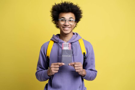 Foto de Attractive smiling African American man holding passport with boarding pass ticket isolated on yellow background. Happy tourist ready for flight. Vacation, travel concept - Imagen libre de derechos
