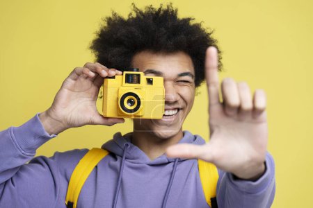 Téléchargez les photos : Jeune photographe afro-américain souriant prenant des photos isolées sur fond jaune. Gros plan portrait de heureux attrayant touriste tenant appareil photo. Vacances, concept de voyage - en image libre de droit