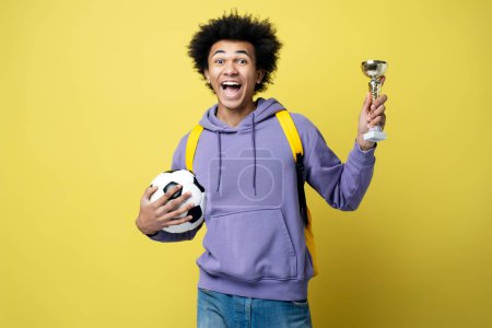 Téléchargez les photos : Young overjoyed African American man football player holding ball and trophy cup, celebration success isolated on yellow background. Sport victory, winning competition concept - en image libre de droit