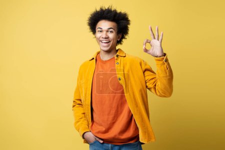 Foto de African American man wearing stylish outfit showing ok sign,  isolated on yellow background - Imagen libre de derechos