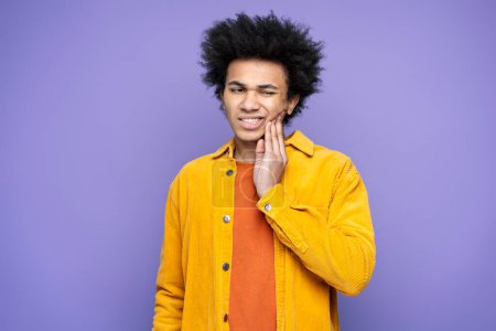 Photo for Young man holding hand on his cheek having a toothache, need medical treatment isolated on purple background. Dental care - Royalty Free Image
